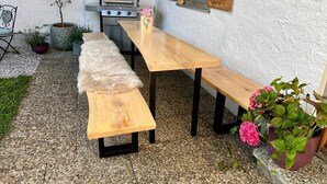 Un patio al aire libre con una mesa y bancos de madera, rodeado de plantas en macetas y una parrilla.