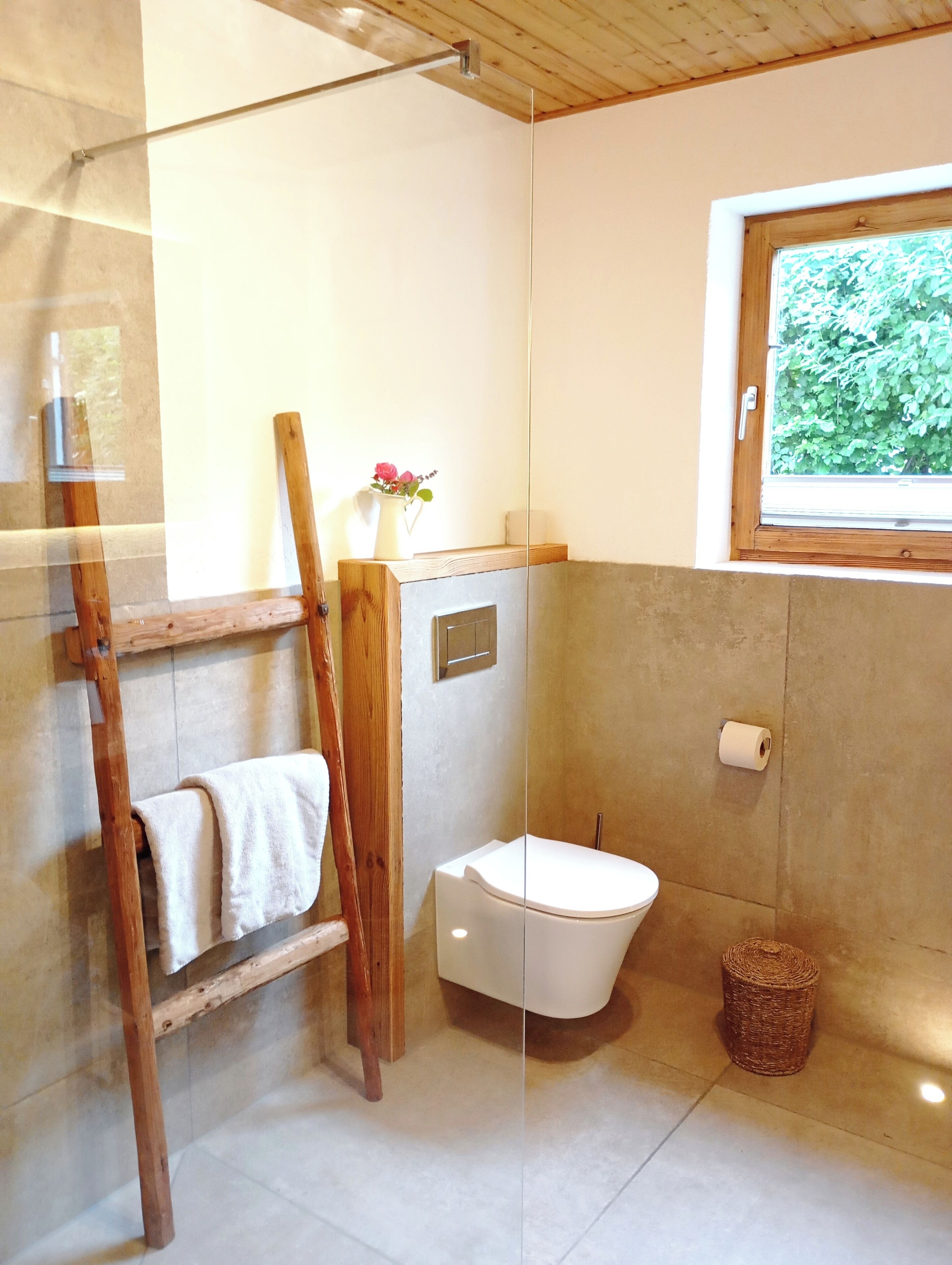 A bathroom with a wooden ladder, a white toilet, a small wicker basket, and a window with a view of greenery.