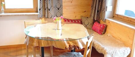 A cozy room with a round wooden table, a glass-top table, a fur rug, wooden chairs, a bench with a fur throw, and a window with curtains.