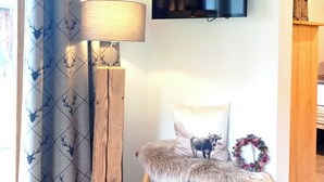 Una habitación con un ventanal grande, una cortina con estampado de ciervo, un televisor montado, un sillón cubierto de piel, una cesta trenzada y una guirnalda colocada en la pared.