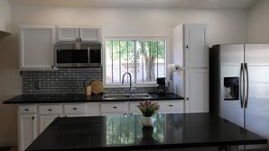 Una cocina moderna con armarios blancos, encimera negra, electrodomésticos de acero inoxidable y un ventanal con vistas a árboles.