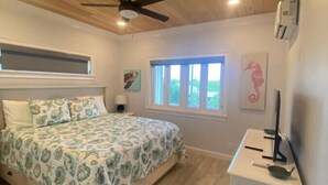 Una camera da letto con un letto, una ventola a soffitto, finestre, opere d'arte e un piccolo armadietto bianco.