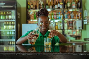 Una persona sirve una bebida en un vaso en un bar, con estantes llenos de botellas al fondo.