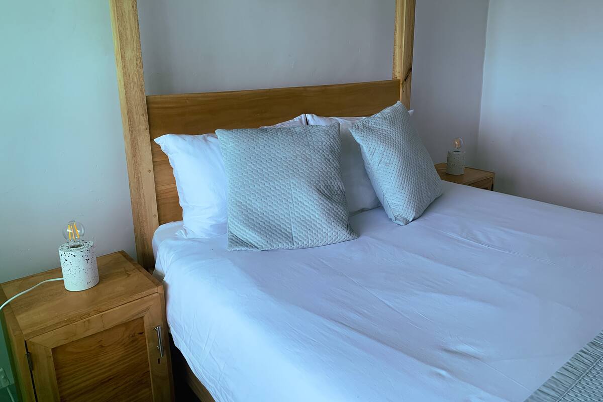 A neatly made bed with white linens and two textured pillows, a wooden headboard, and a small bedside table with a lamp and a decorative item.