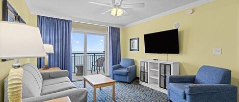 A hotel room with a ceiling fan, a flat-screen TV, a sofa, a chair, a small table, and a view through a glass door to the outside.