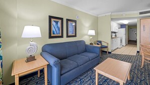 A hotel room with a blue sofa, a wooden table, a small side table with a lamp, and a kitchenette in the background.