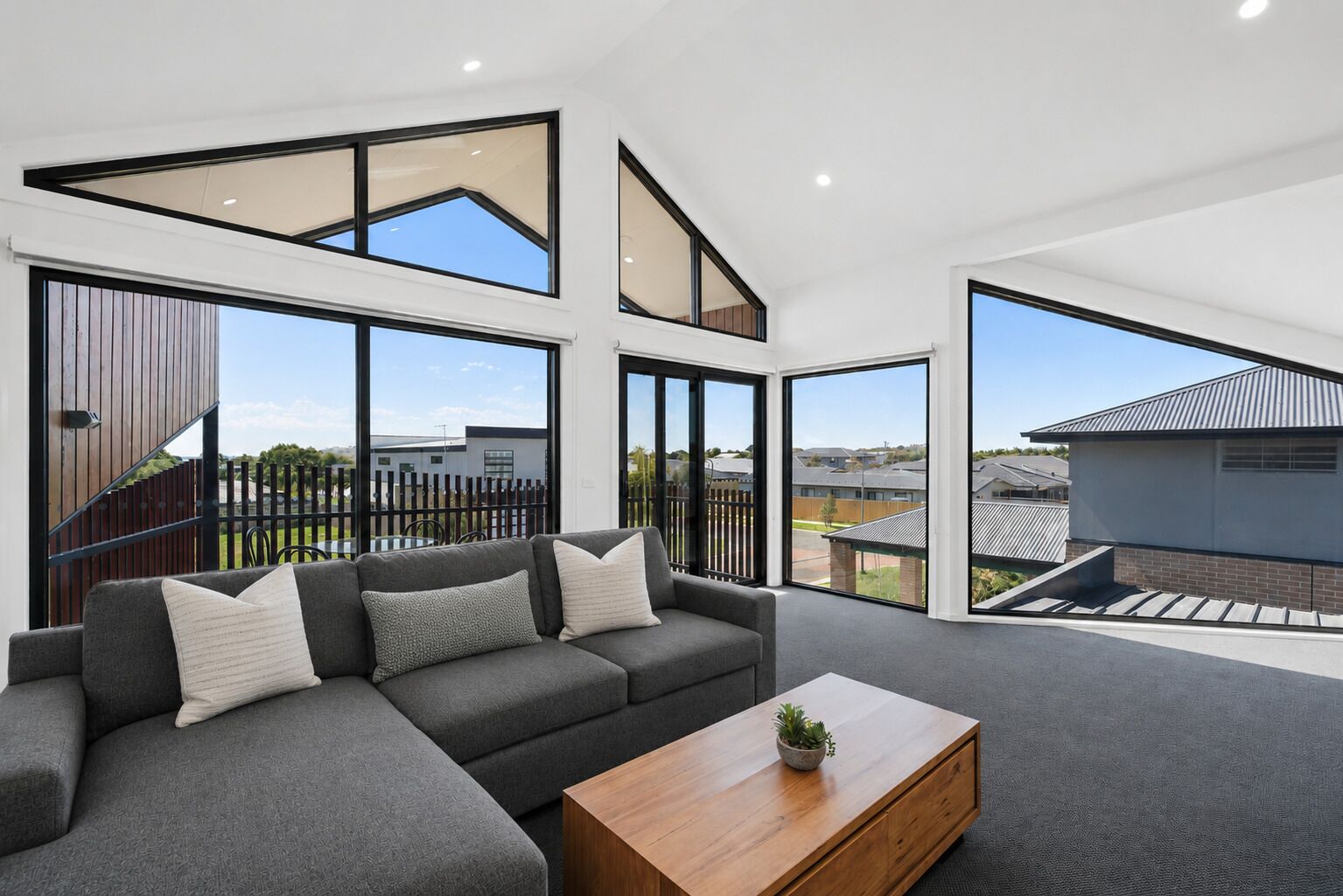 A modern living room with a sectional sofa, a wooden coffee table, and large windows offering a view of the neighborhood.