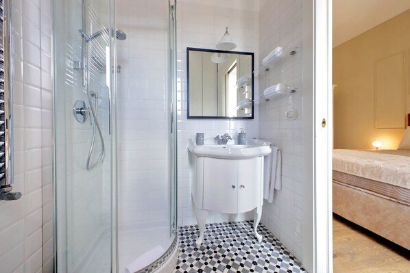 A bathroom with a glass shower enclosure, a white sink with a mirror above it, and a bed with a beige mattress visible in the adjacent room. - Amazing penthouse in the heart of Rome (Roma)