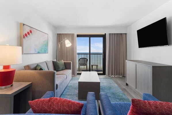Una habitación de hotel moderna con sofá, una mesa de centro, televisor y balcón con vistas al mar.