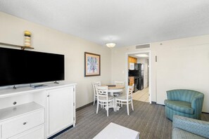 A hotel room with a flat-screen TV, a dining table with chairs, a blue armchair, and a kitchenette in the background.