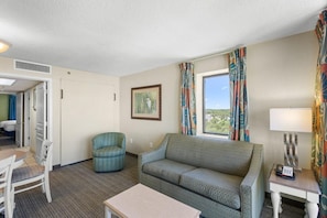 Et hotelværelse med sofa, lænestol, bord og et vindue med gardiner.