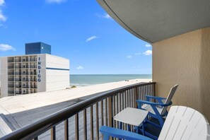 A balcony with a view of the beach, a building with 'INDIGO' signage, and a railing.