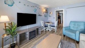 A living room with a flat-screen TV, a dining table with chairs, a blue sofa, and a potted plant.