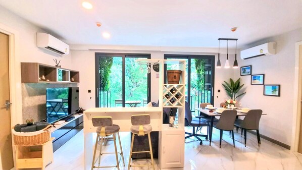 A modern kitchen with a dining area, large windows, and a view of greenery.