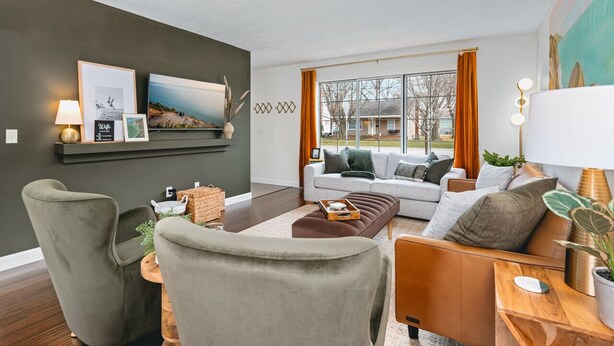 Een moderne woonkamer met een grijze bank, een bruin voetenbankje en een houten bijzettafeltje. Er hangen afbeeldingen in lijsten aan de muur en er is een raam met gordijnen.