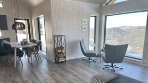 Een ruime woonkamer met een houten vloer, een eettafel met stoelen, een grijze bank en een groot raam met uitzicht op een bergachtig landschap.