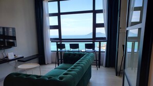 A modern room with a green sofa, a small table, and a TV. A large window offers a view of the sea and a distant island. - Jesselton Quay 23A Sunset Seaview  (Kota Kinabalu)