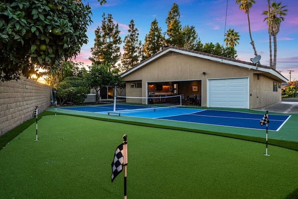 A tennis court with a net, surrounded by a green lawn and a building with a garage. - Pickleball | Putting Green | 5 BR | 3 mi to Disney (Anaheim)