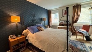 Une chambre à coucher avec un grand lit, une table de chevet, un bureau, une chaise et une fenêtre avec des rideaux.