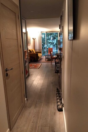 A hallway leading to a balcony with a view of trees and a building. There is a door on the left, a chair and a table on the right, and a rug on the floor.