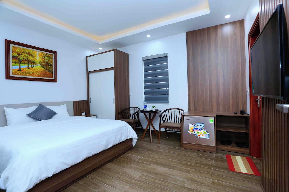 A hotel room with a bed, a desk, a chair, a small refrigerator, and a window with blinds.