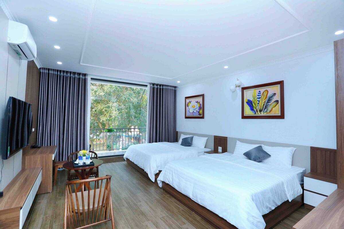 A hotel room with two beds, a desk with a chair, a TV, and a balcony with curtains.