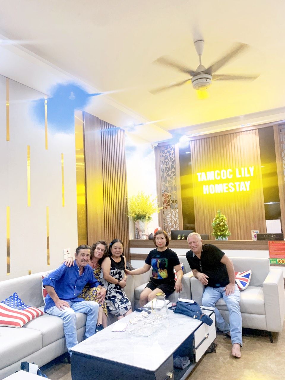A group of people seated on a sofa in a room with a ceiling fan, a coffee table, and a wall with a sign that reads 'TAMCOC LILY HOMESTAY'.