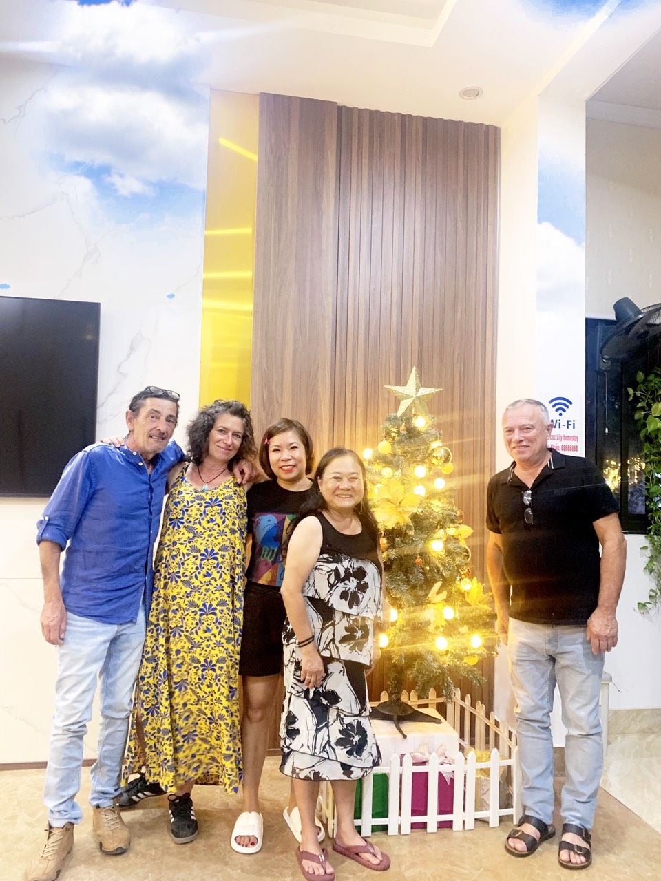 Un grupo de personas de pie frente a un árbol de Navidad con una estrella en la punta, en una habitación con pared de paneles de madera y un televisor de pantalla plana montado en la pared.