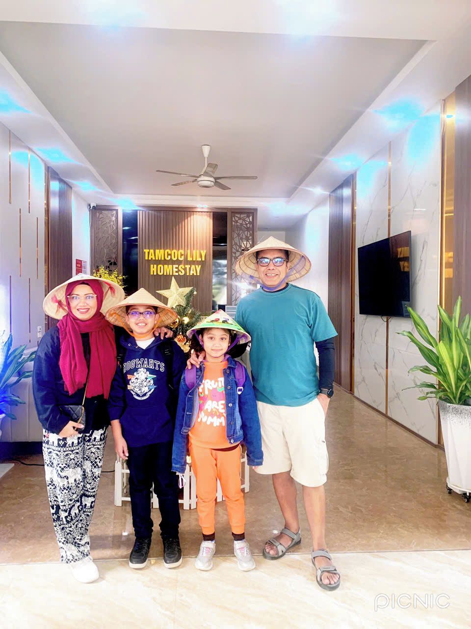 A hotel lobby with a modern design, a ceiling fan, a sign reading "TAMCOC LILY HOMESTAY", and a large potted plant.