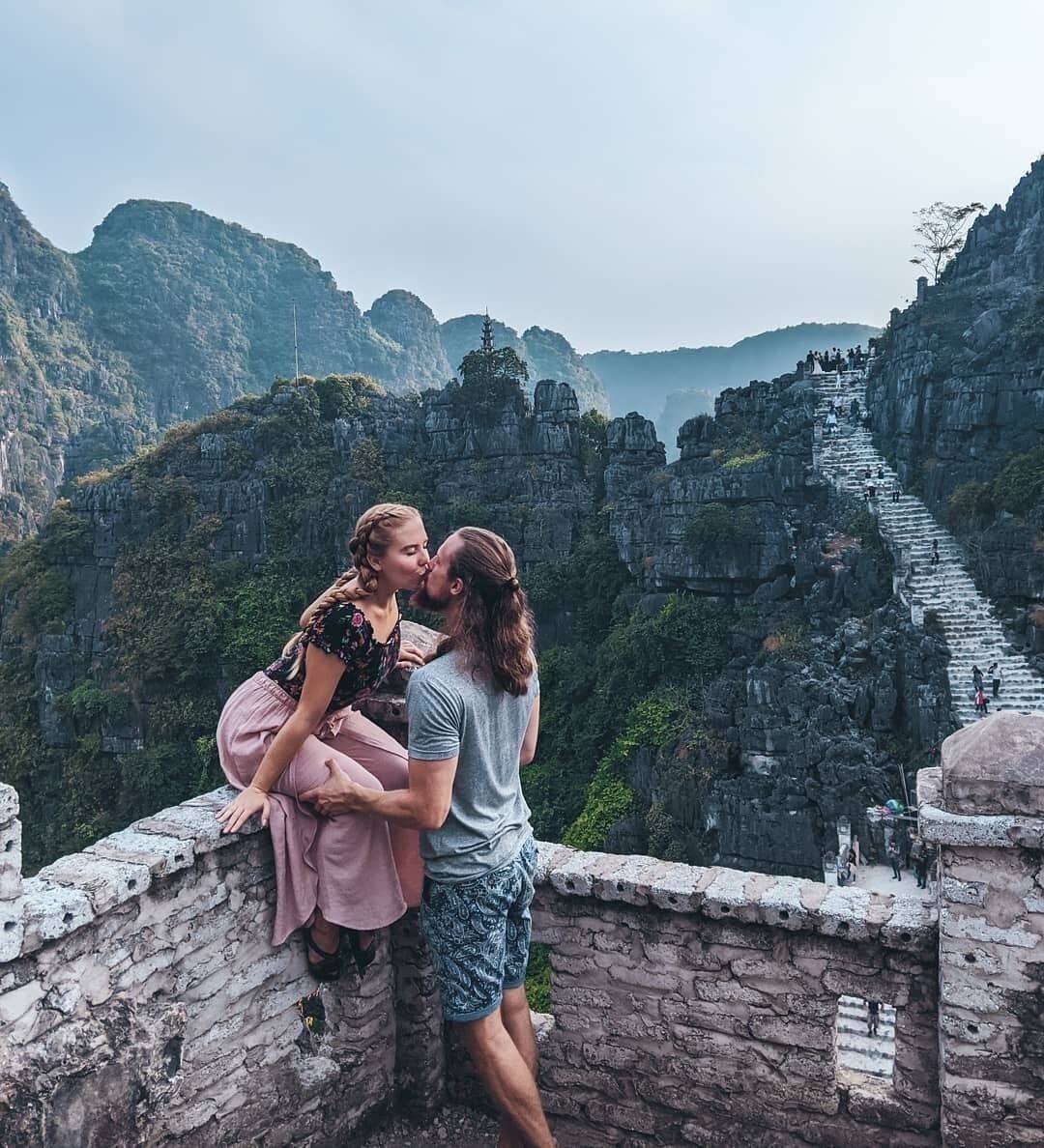 A couple kissing on a stone ledge overlooking a mountainous landscape with a stone staircase.
