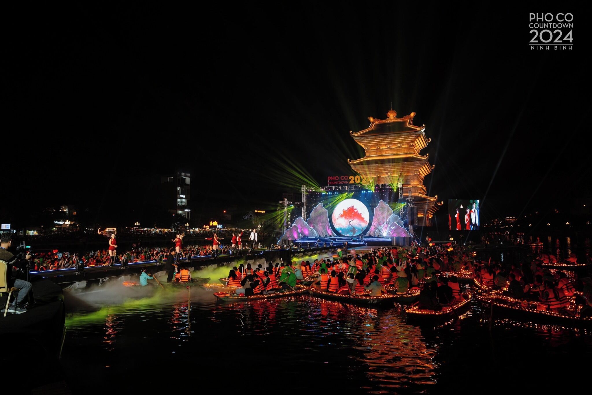 Vista nocturna de una pagoda tradicional con un escenario frente a ella, iluminada por focos y rodeada por una multitud en barcos.