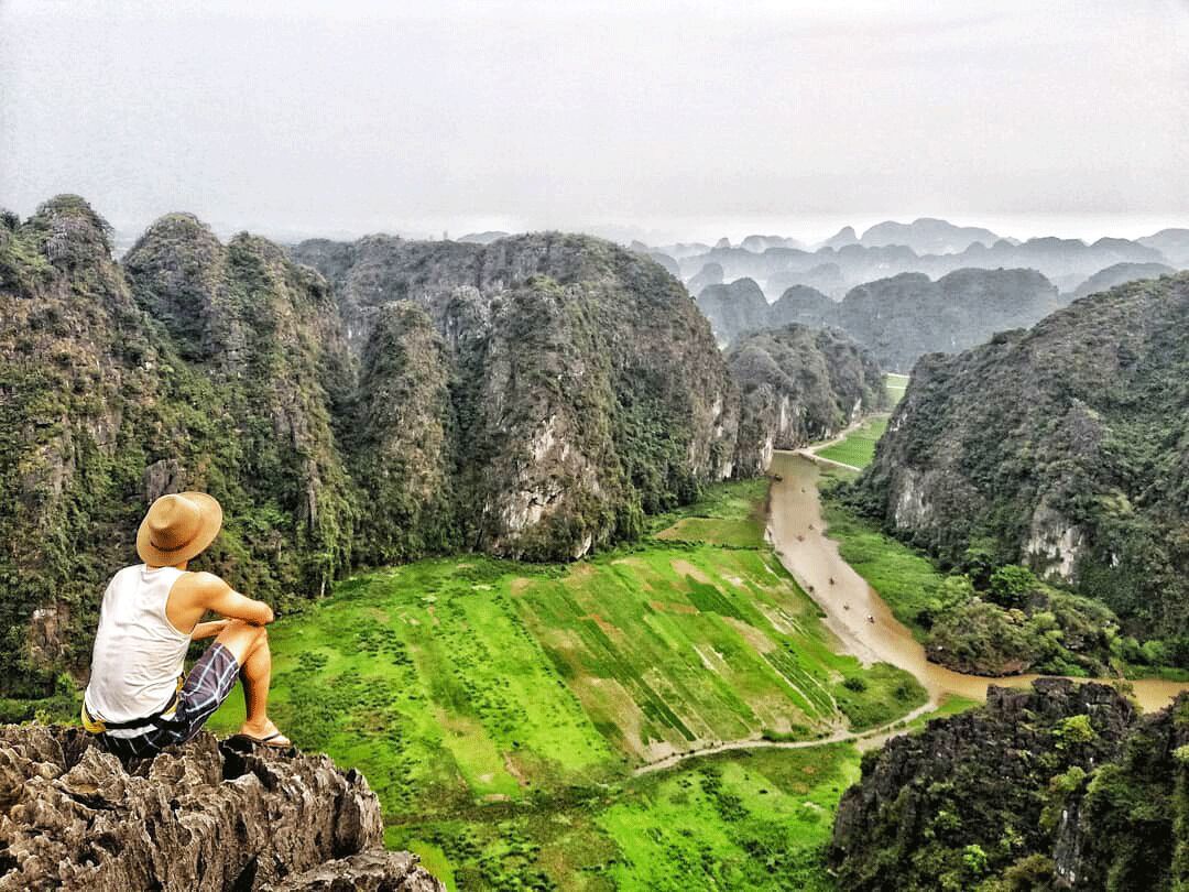 A person sitting on a rock overlooking a valley with a river and green fields.