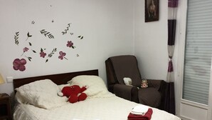 Un lit bien fait, avec des oreillers et une couverture, une chaise sur laquelle est posé un ours en peluche, et un mur décoré de motifs floraux.