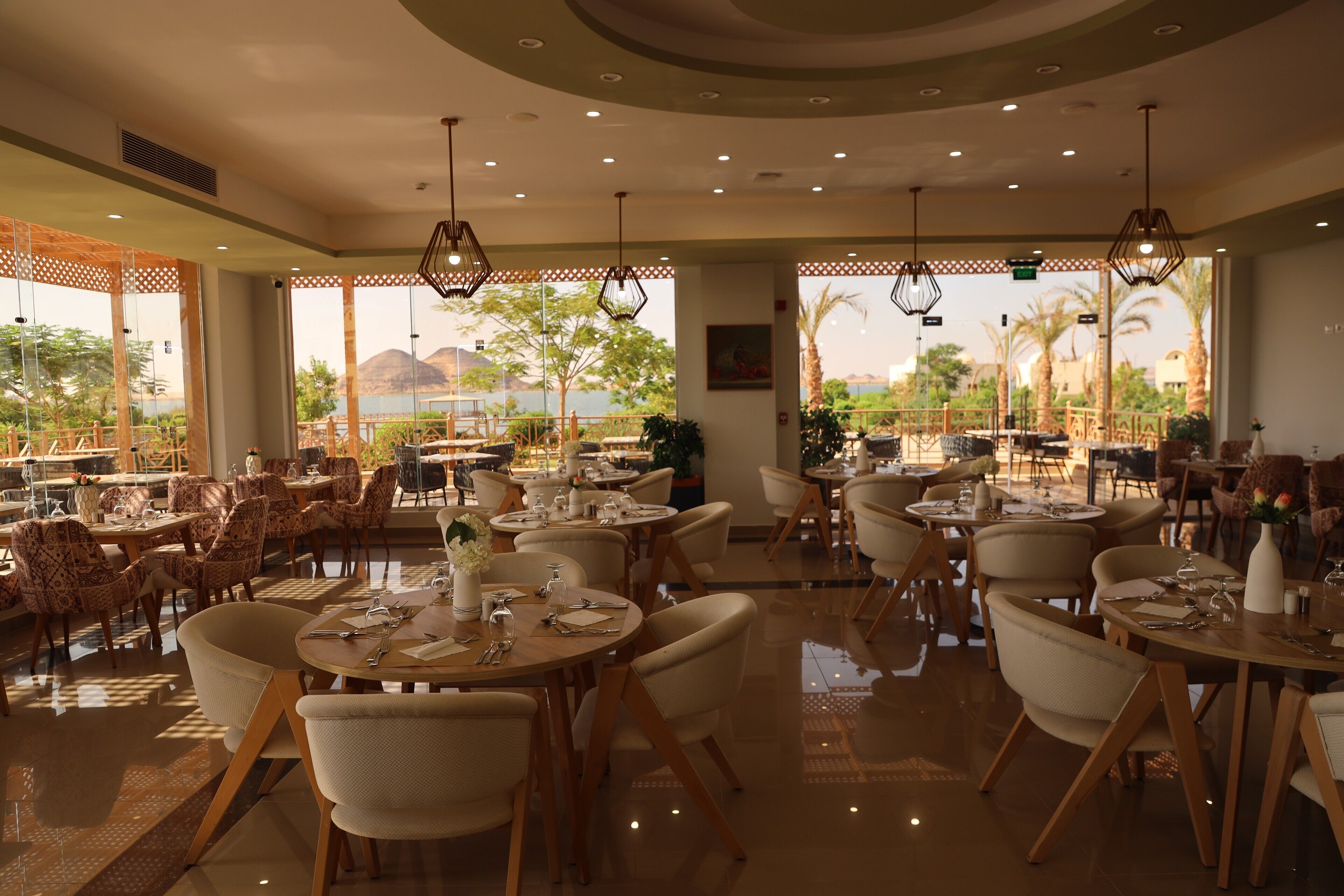 Un restaurante espacioso con numerosas mesas y sillas, amplias ventanas que ofrecen vistas a palmeras y montañas, e iluminación colgante moderna.