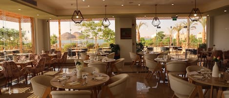 Un restaurante espacioso con numerosas mesas y sillas, amplias ventanas que ofrecen vistas a palmeras y montañas, e iluminación colgante moderna.