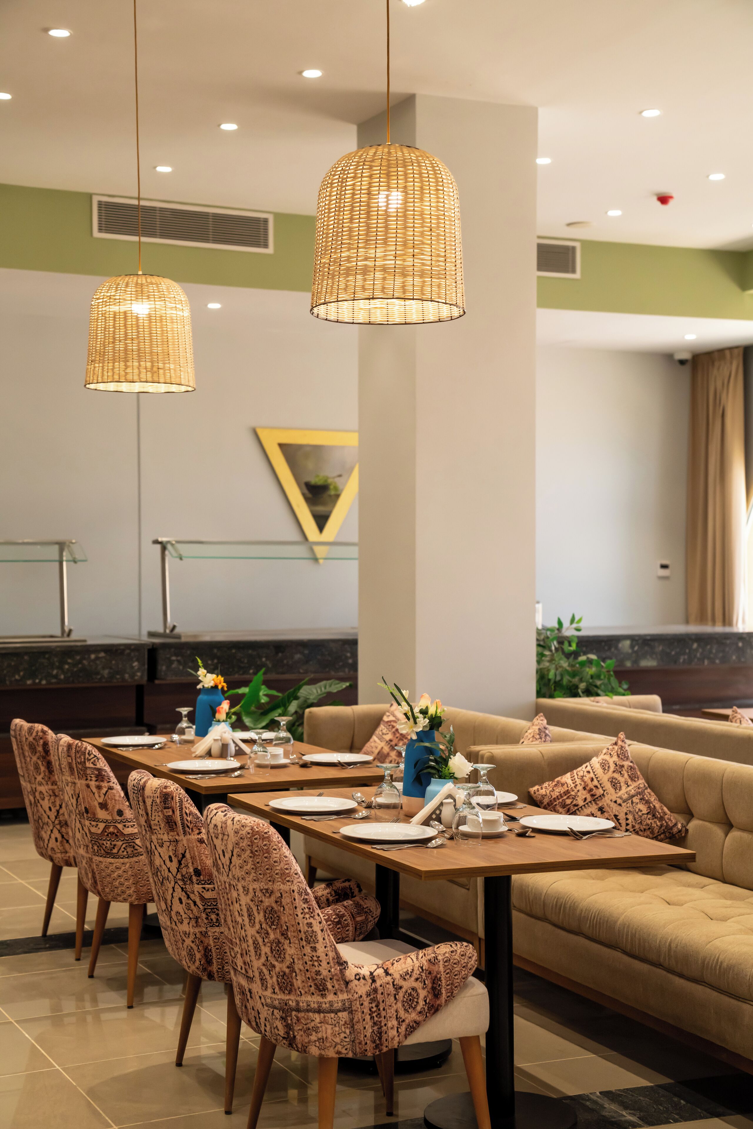 A dining area with a long table set for four, surrounded by patterned chairs, a sofa, and two hanging wicker lamps.