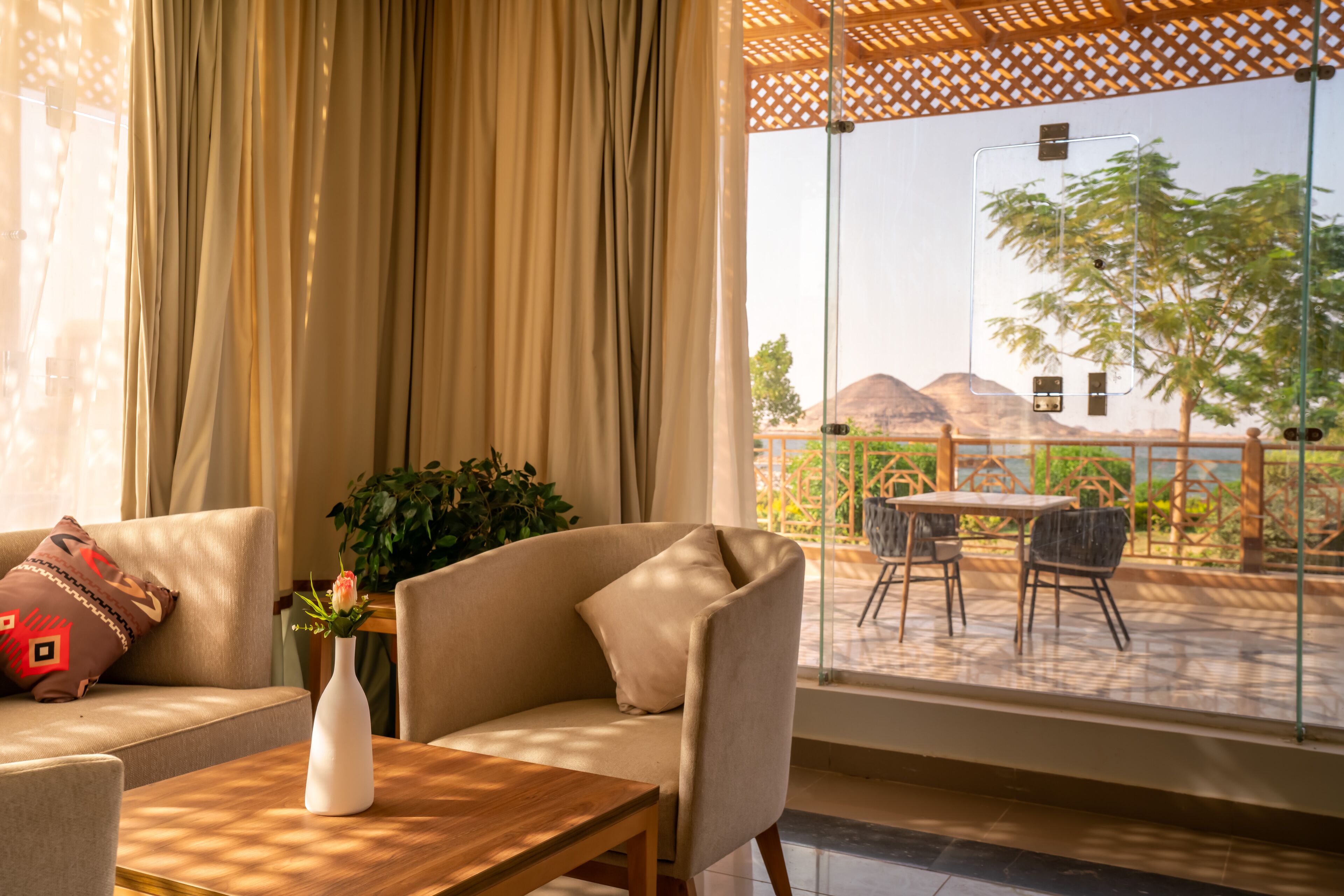 A room with a sofa, a table, and a view of a terrace with outdoor furniture and a mountain in the background.