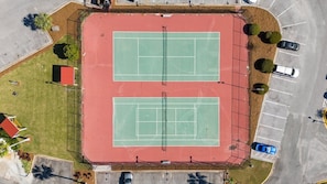 An aerial view of a tennis court complex with two courts, parking area, and surrounding landscaping.