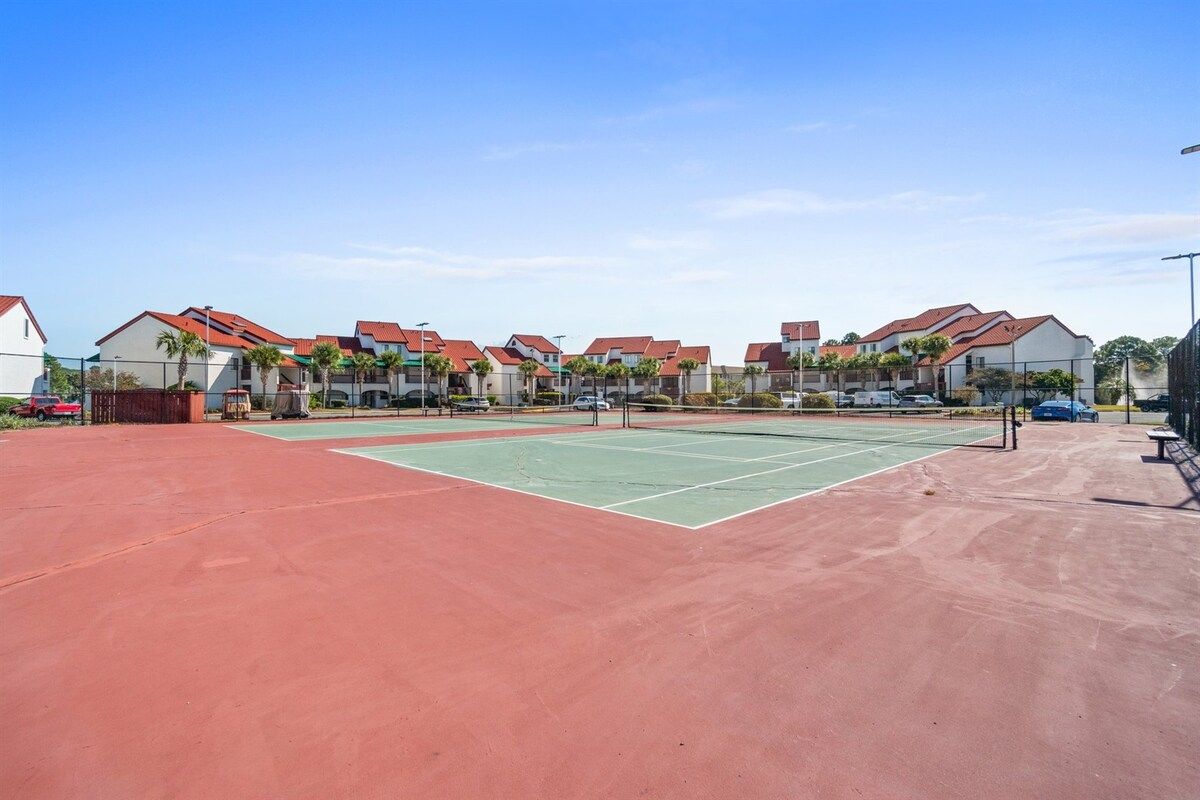 Een tennisbaan met een rode ondergrond, omgeven door woongebouwen met rode daken.