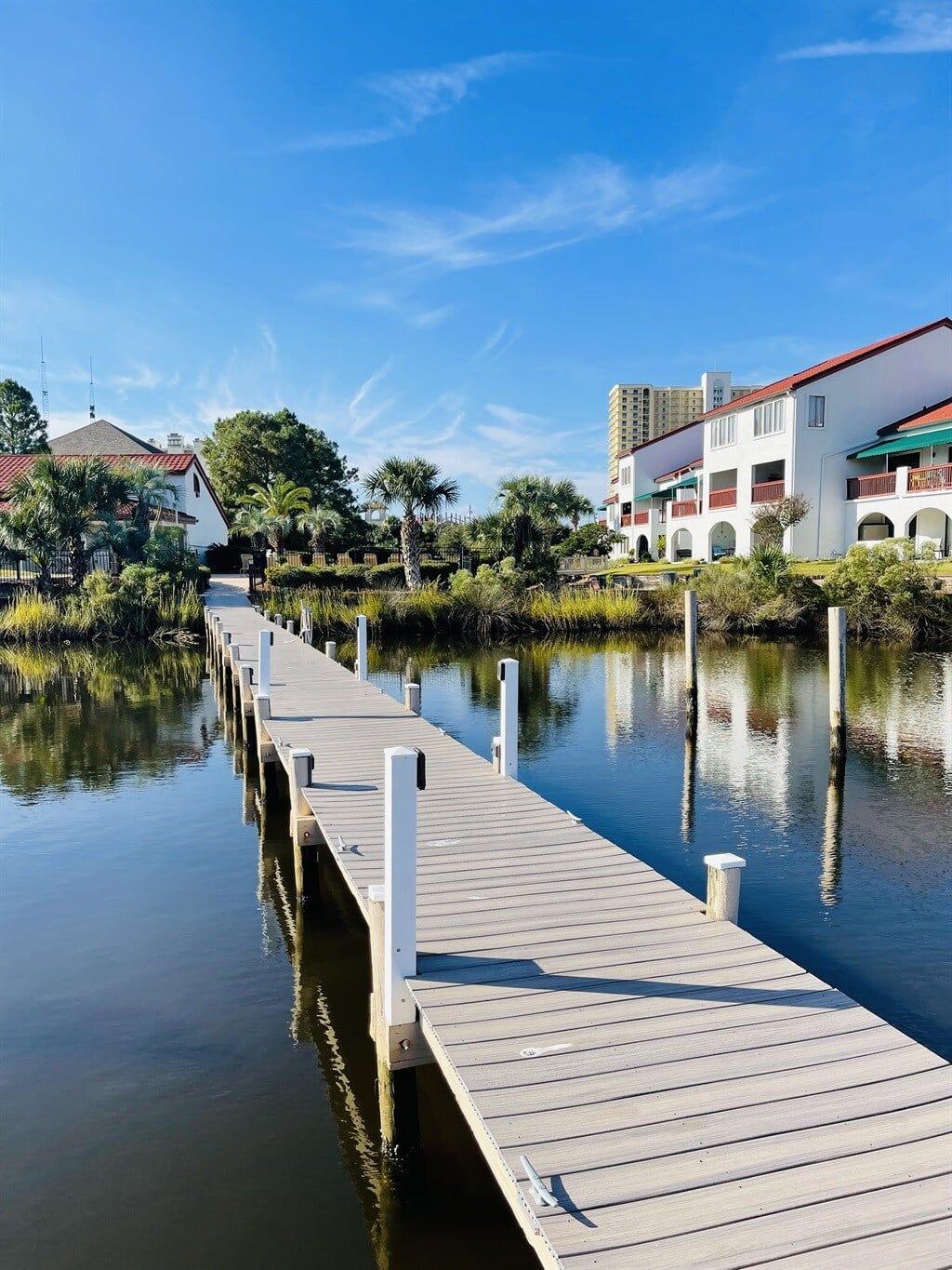 A wooden pier extending into calm water, surrounded by greenery and buildings with red roofs.