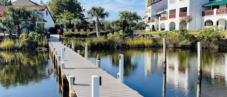A wooden pier extending into calm water, surrounded by greenery and buildings with red roofs.