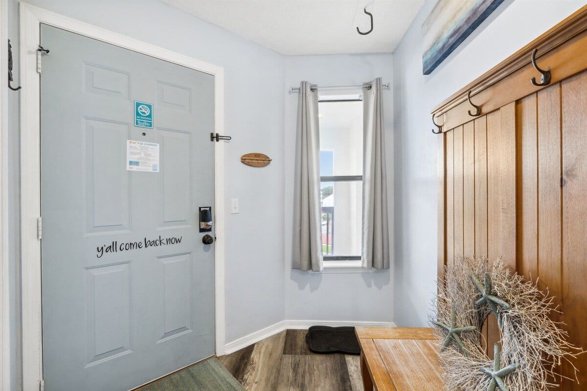 A room with a door marked 'y'all come back now,' a window with curtains, a bench, and a wall-mounted coat rack.