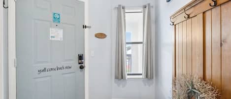 Una habitación con una puerta que dice 'y'all come back now', una ventana con cortinas, un banco y un perchero de pared.