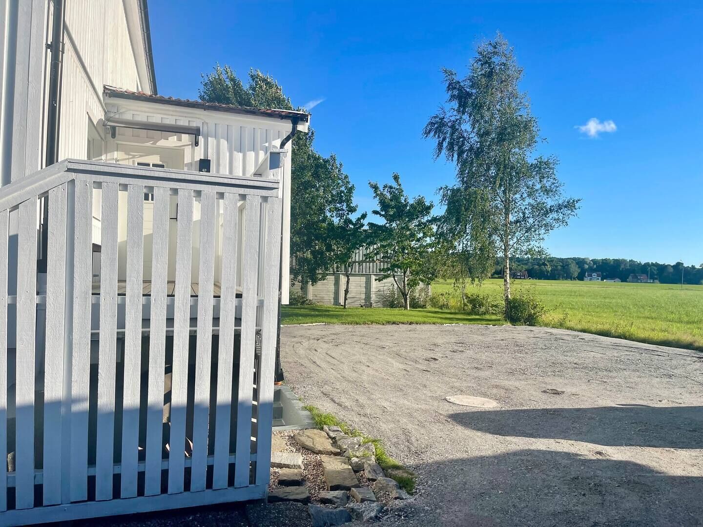 A white building with a wooden fence, a gravel driveway, and a grassy field with trees in the background.