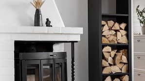 A fireplace with a white mantel and black surround, a black wood-burning stove, and a tall black bookshelf filled with neatly stacked firewood.