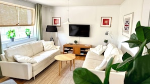 A modern living room with a white sofa, a coffee table, and a TV. There are two potted plants, a lamp, and framed pictures on the wall.
