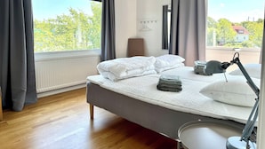A modern bedroom with a large bed, a bedside table, a lamp, and a view of trees and a house through the window.