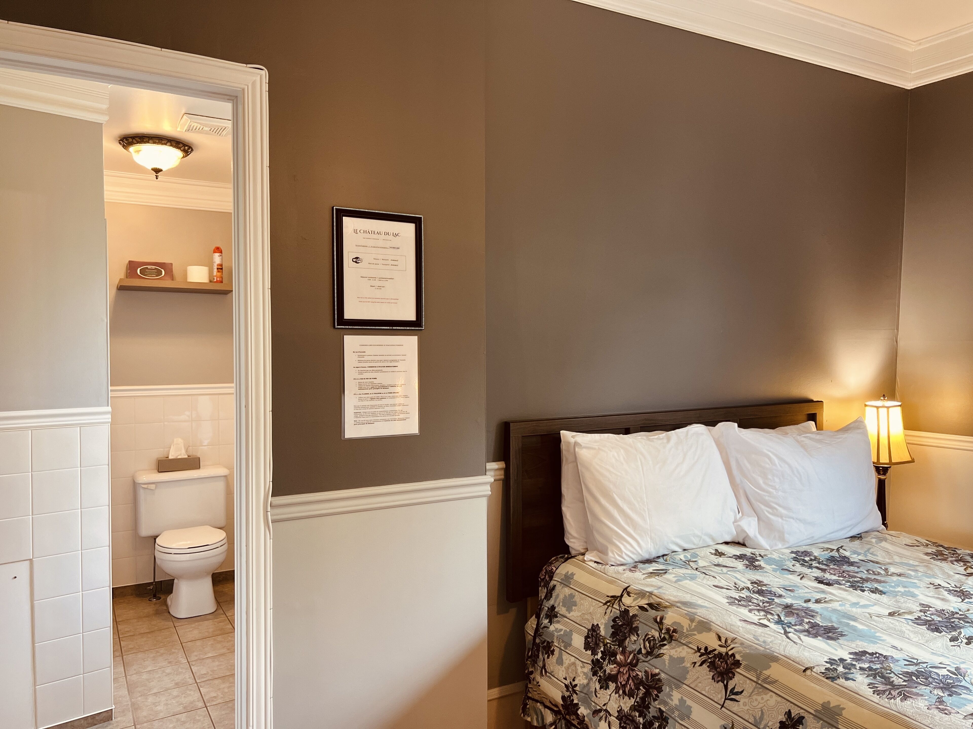 A hotel room with a bed, a nightstand, a wall-mounted lamp, a framed certificate, and a bathroom visible through an open door.