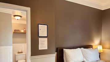 A hotel room with a bed, a nightstand, a wall-mounted lamp, a framed certificate, and a bathroom visible through an open door.