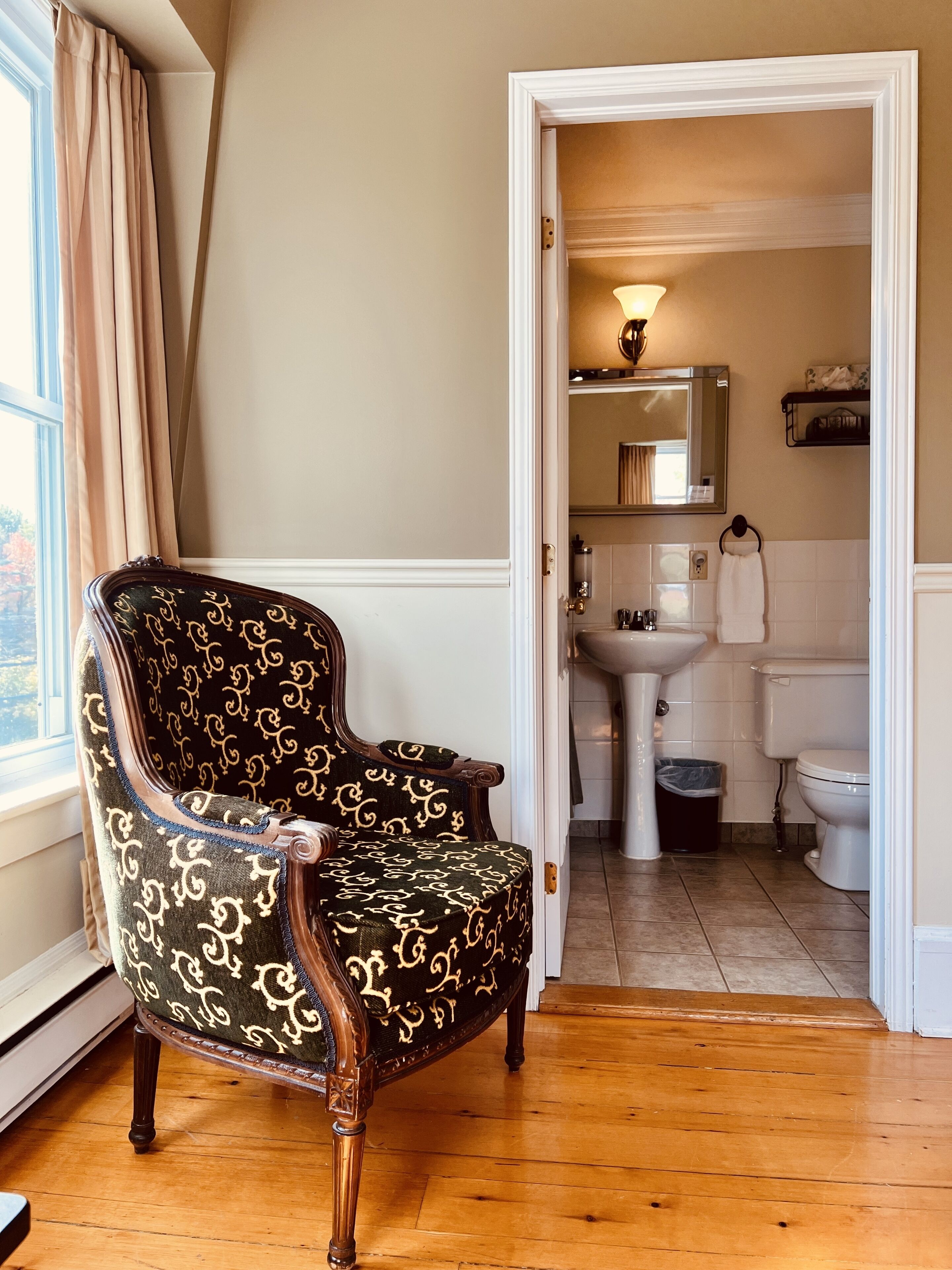 A room with a wooden floor, a patterned armchair, a doorway leading to a bathroom with a sink, toilet, and mirror.